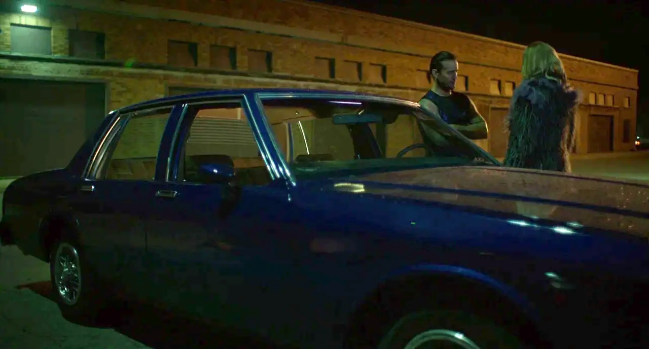Rabid (2019) movie still: wide — A dark parking lot at night features a large, brick building in the background. A man and woman stand next to a…
