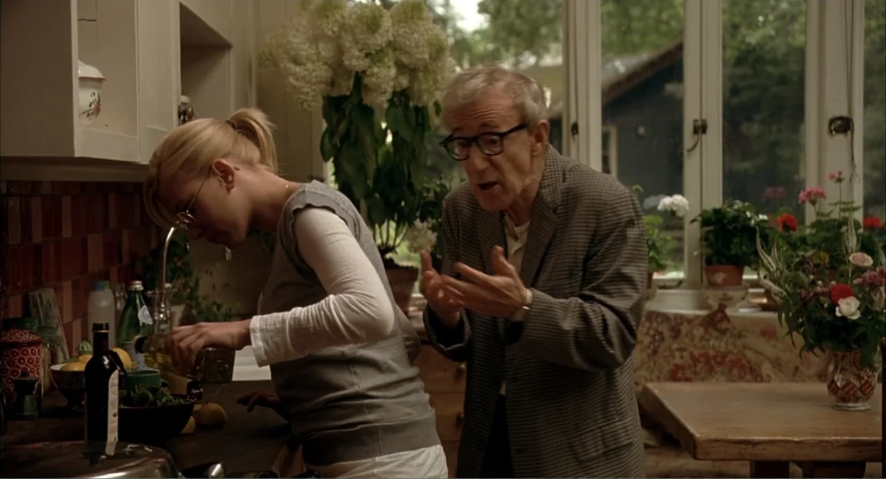 Scoop (2024) movie still: medium — A well-lit kitchen with wooden cabinetry, potted hydrangea plants, and a large windowed wall. An older man…