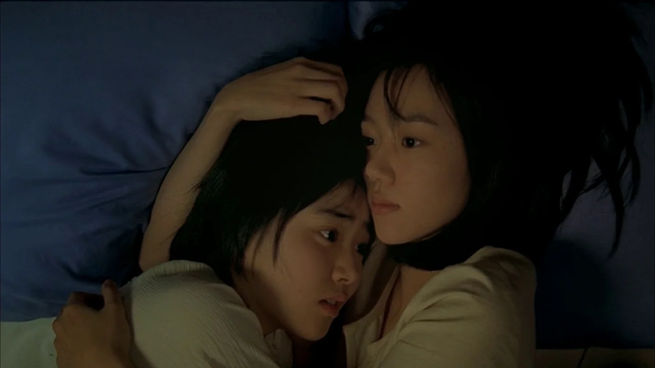 A Tale of Two Sisters (2003) movie still: closeup — Two individuals lie together on a dark blue surface. They are embraced while lying down in a dim…