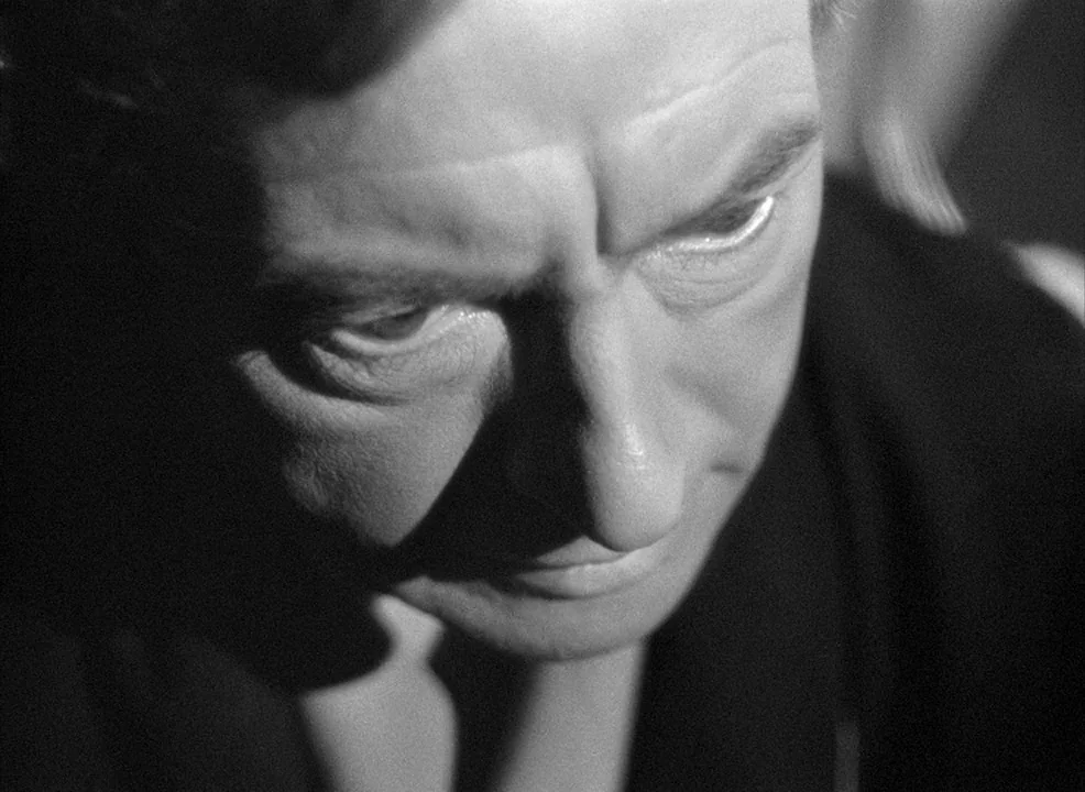 Notorious (1946) movie still: closeup — A dark indoor space defined by high-contrast black and white lighting. A man looks downward with a furrowed…