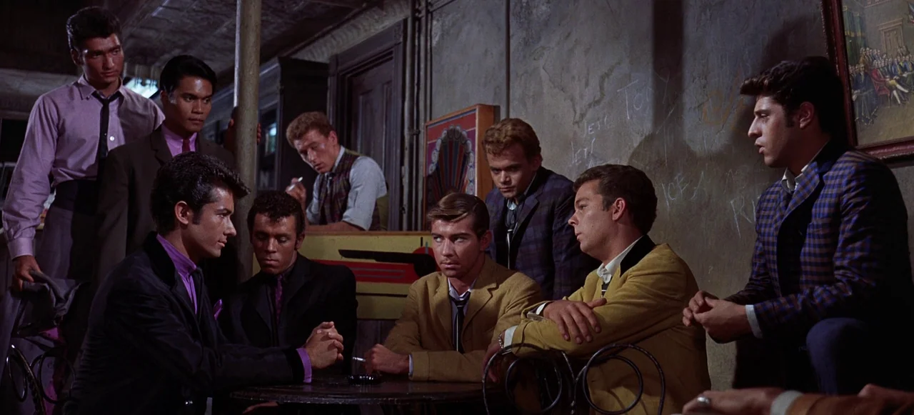 West Side Story (1961) movie still: medium — A dimly lit room with distressed plaster walls marked with graffiti and dark woodwork. Several young…