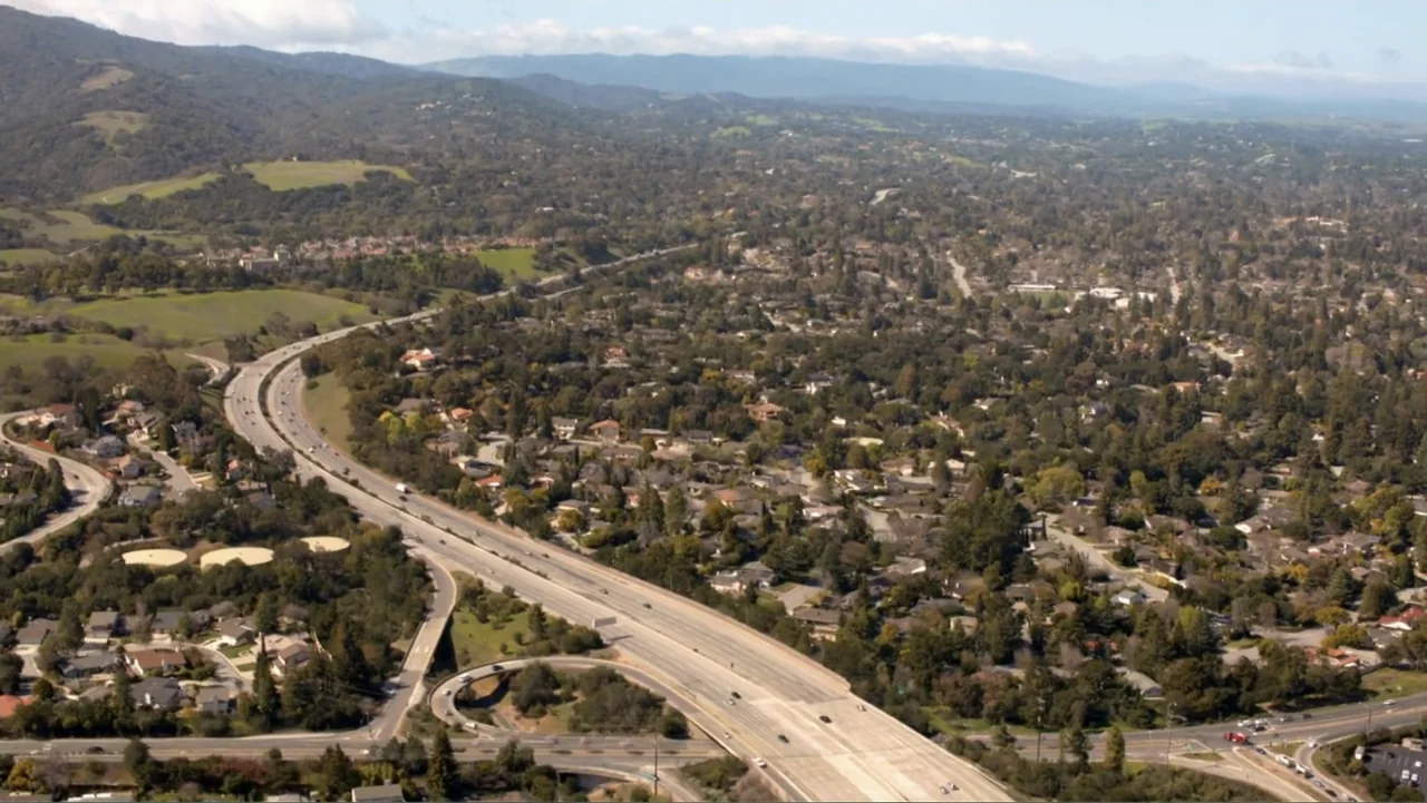 Clear History (2013) movie still: establishing — A suburban landscape features a sprawling highway interchange surrounded by dense green trees.…