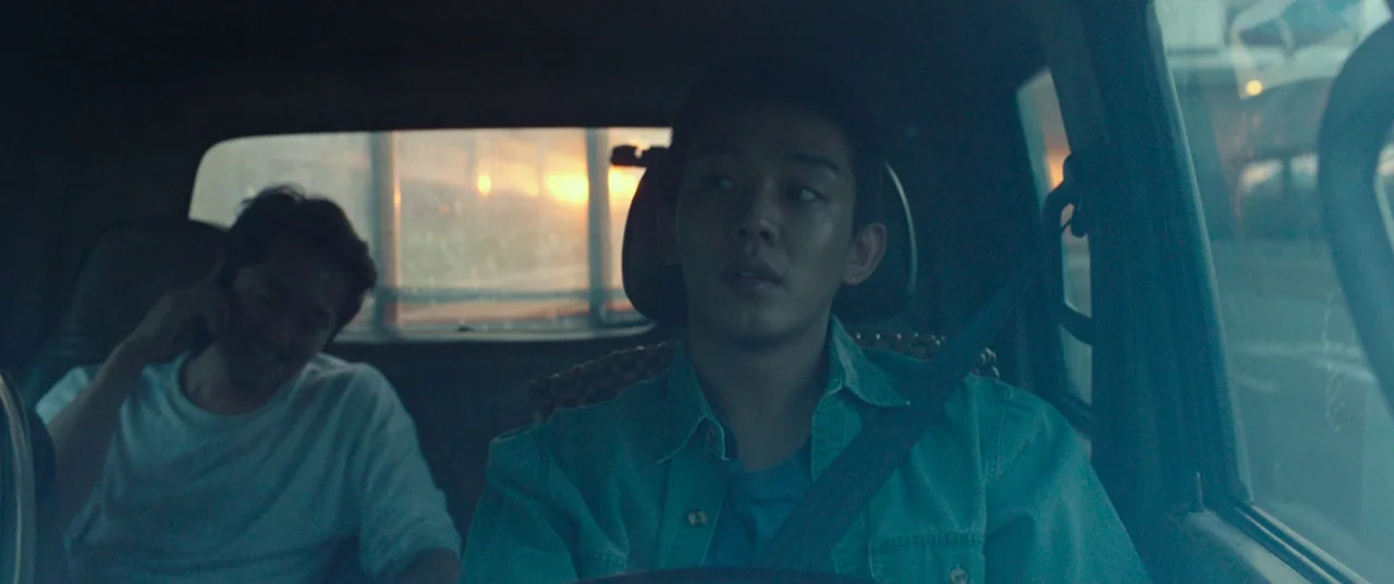 Burning (2018) movie still: medium — The interior of a moving car during sunset with a driver and rear passenger. The driver looks forward while the…