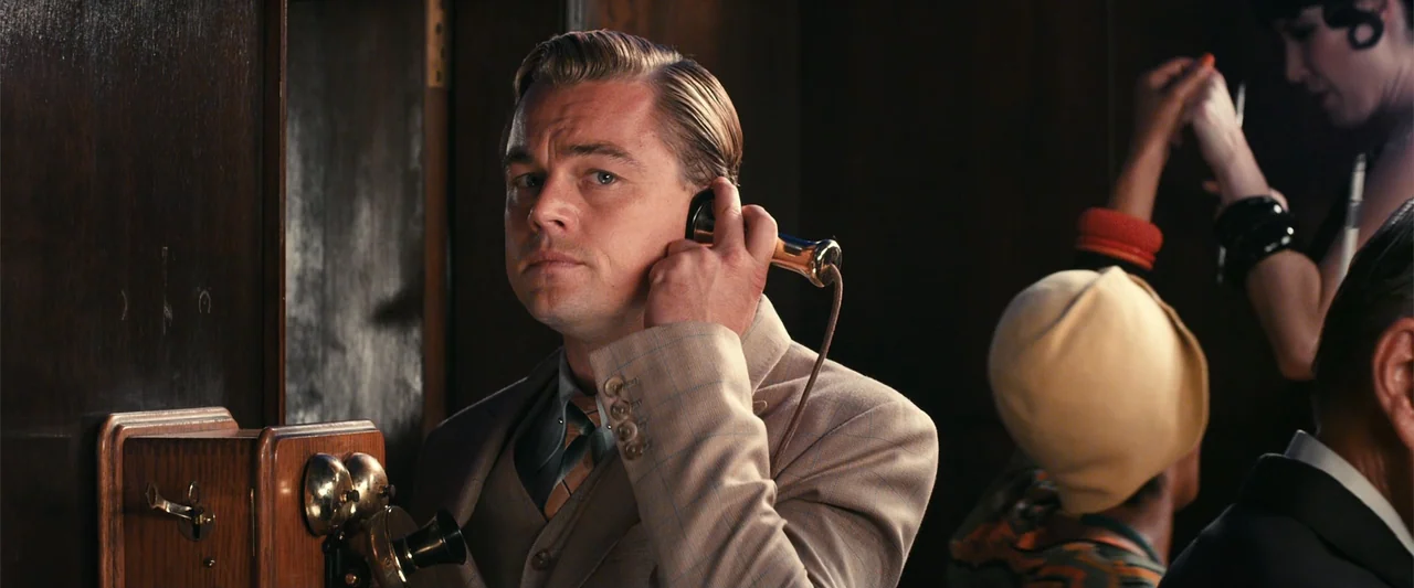 The Great Gatsby (2013) movie still: medium — A wood-paneled room interior from the 1920s with an antique telephone mounted on the wall. A man in a…