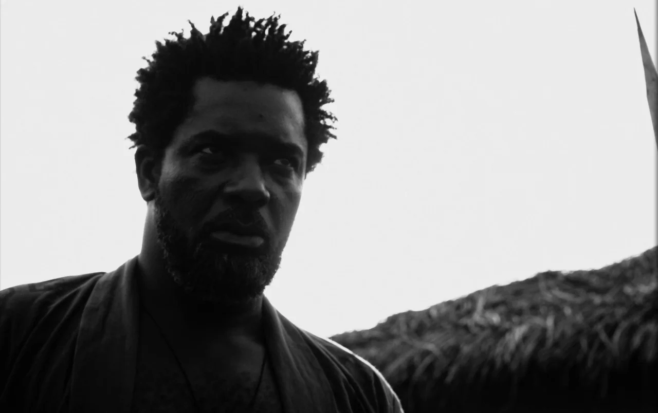 Mami Wata (2023) movie still: closeup — A man stands outdoors under a bright sky next to a thatched roof structure. He looks away from the camera…