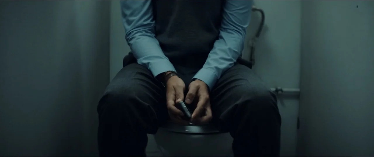 Bonus Track (2024) movie still: medium — A cramped, dimly lit toilet cubicle with plain grey walls and a ceramic toilet. A man wearing a sweater…