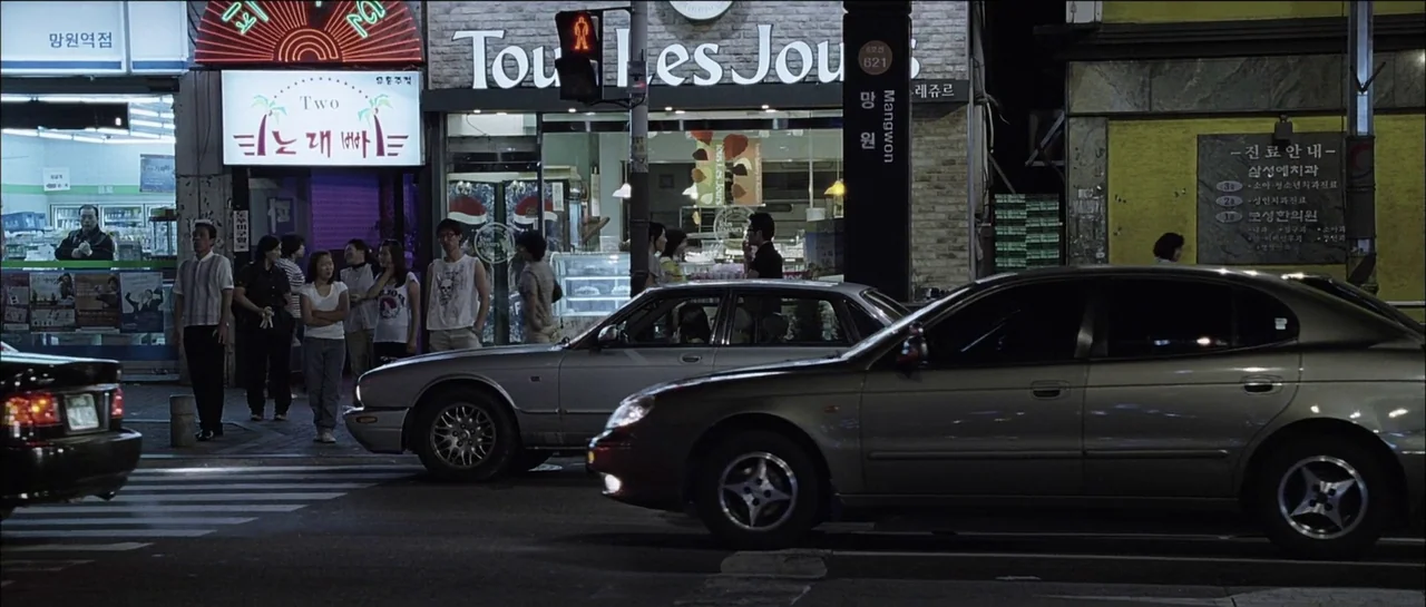 The Chaser (2008) movie still: wide — A Korean urban street corner at night with storefronts and passing vehicles. People stand on the sidewalk near…