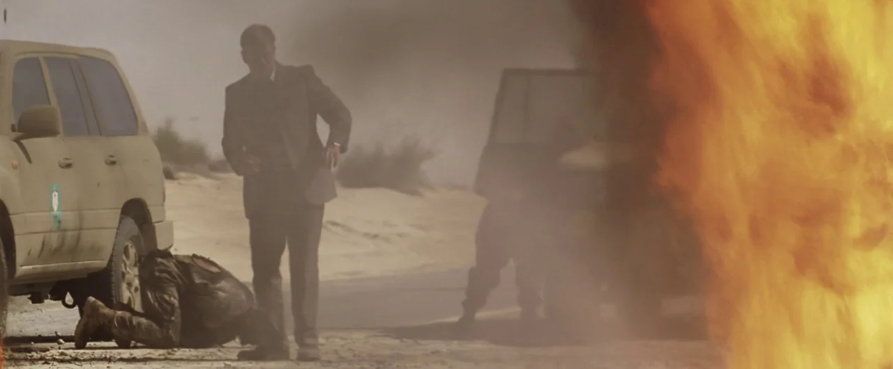 Syriana (2005) movie still: wide — A sandy desert road features a large explosion and two parked SUVs. One man stands by a car while another figure…