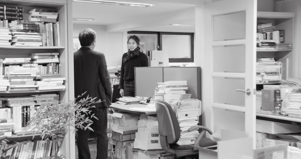 The Day After movie still: medium — A cluttered office filled with tall shelves packed with numerous books and paperwork. A man in a blazer stands…