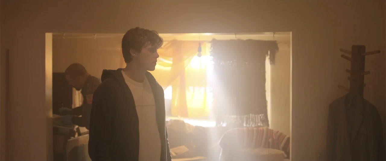 Trap House (2025) movie still: medium — A hazy room with sun shining through a window behind sheer yellow curtains. A young man stands in the…