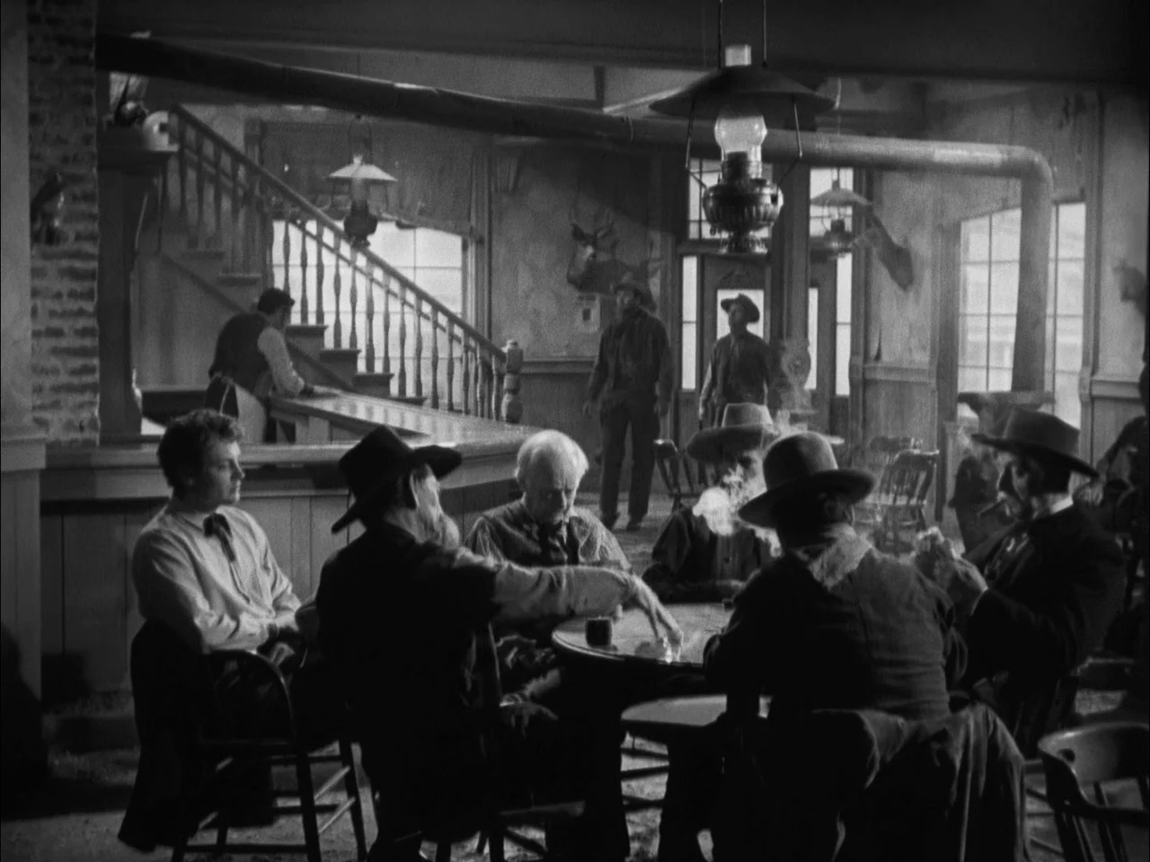 The Ox-Bow Incident (1942) movie still: wide — A dimly lit 19th-century saloon features a wooden bar, staircase, and game tables. Several men sit…