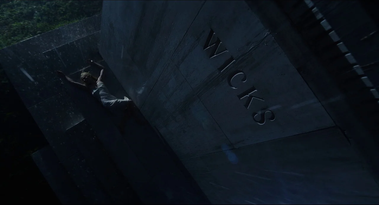 Wake Up Dead Man (2025) movie still: wide — A dark, rain-slicked granite tombstone with the name WICKS engraved on its face. A young woman lies…