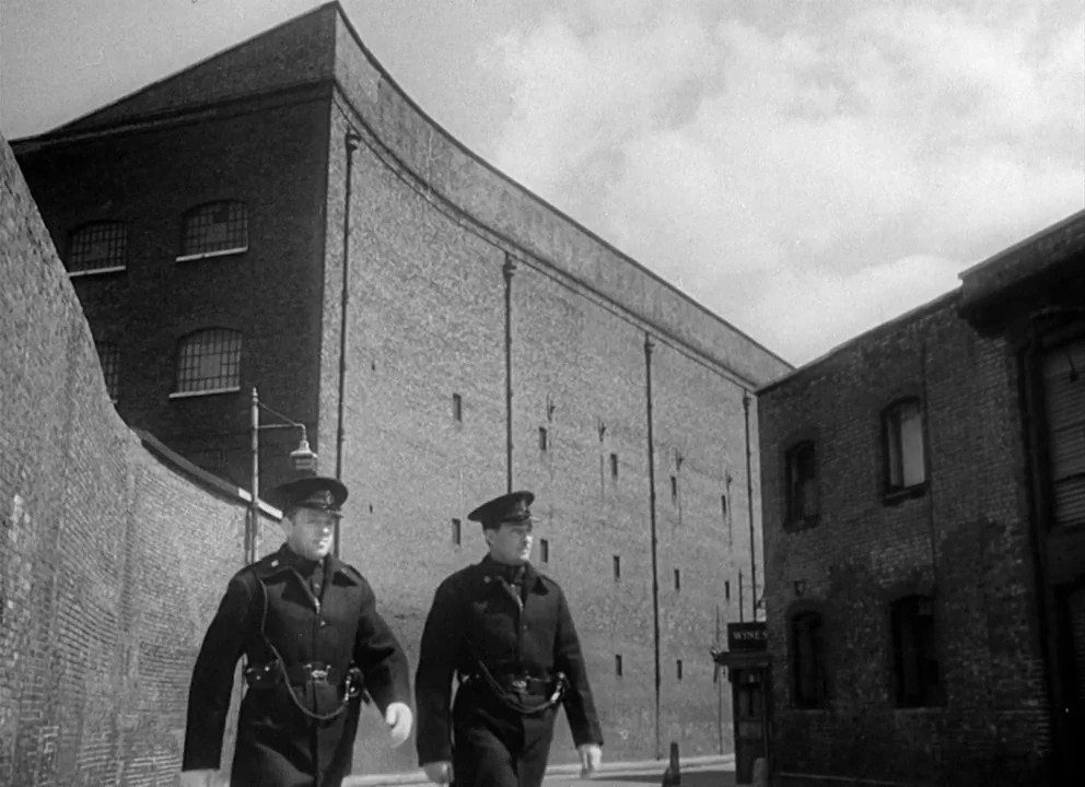 Odd Man Out (1947) movie still: fullbody — An industrial alleyway framed by tall, multi-story brick warehouse buildings with many small windows. Two…