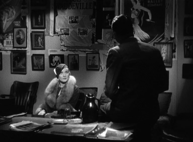 Blonde Venus (1932) movie still: medium — An office with walls covered in numerous framed posters and pictures. A woman sits behind a desk while a…