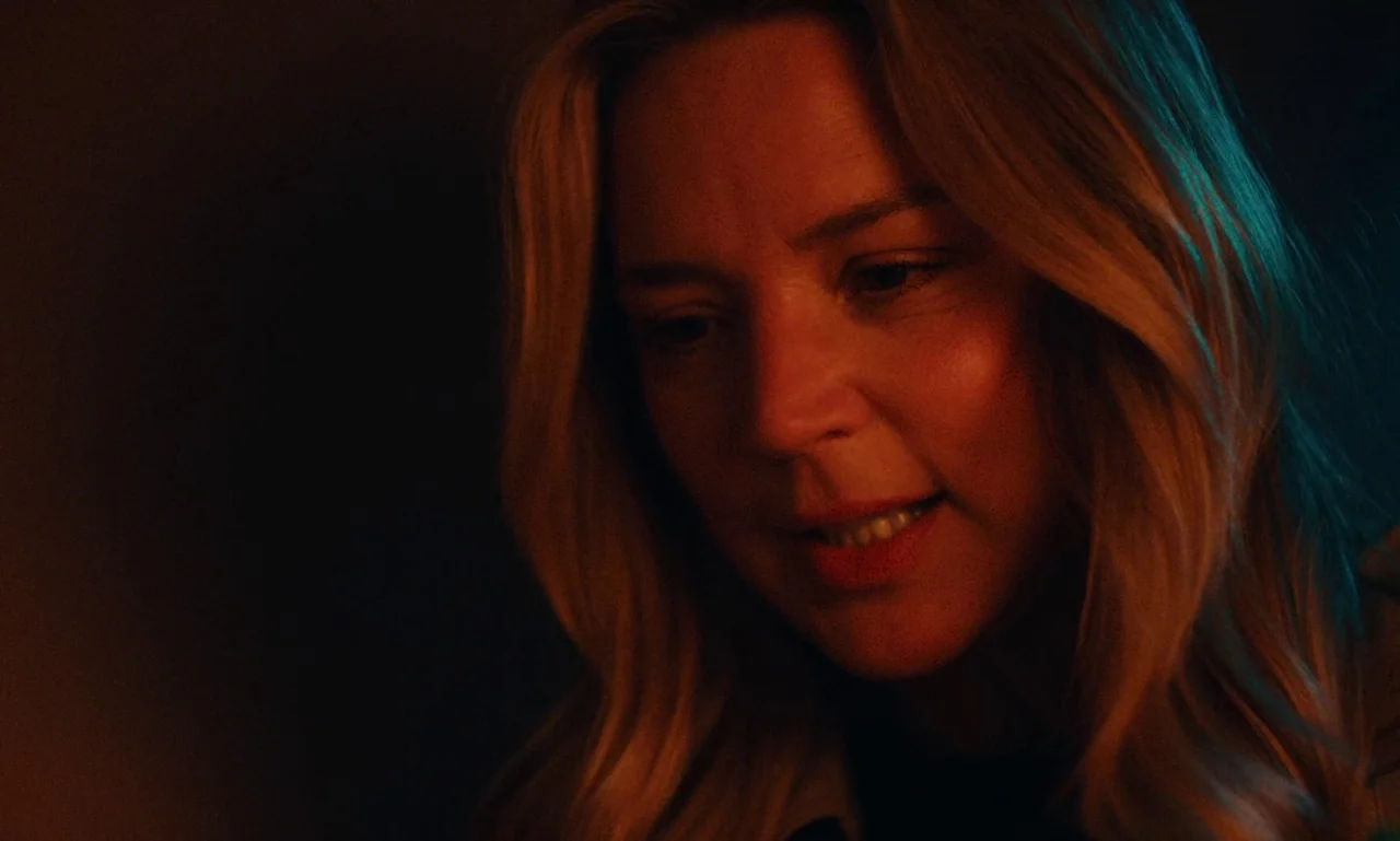 Just The Two Of Us (2023) movie still: closeup — A dimly lit room with heavy shadows obscuring the background. A woman looks downward while…