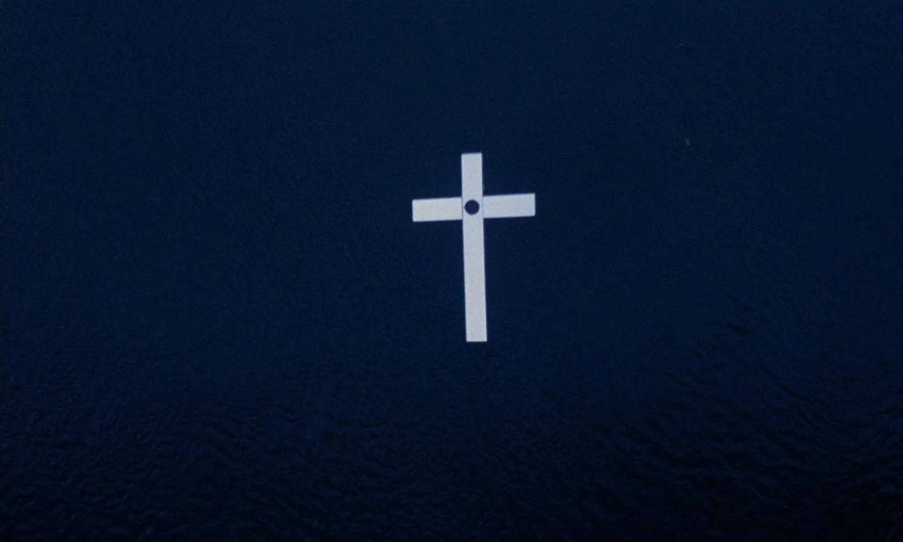 The Church (1989) movie still: closeup — A white cross floats on the surface of dark, rippling deep blue water. The water texture creates subtle…