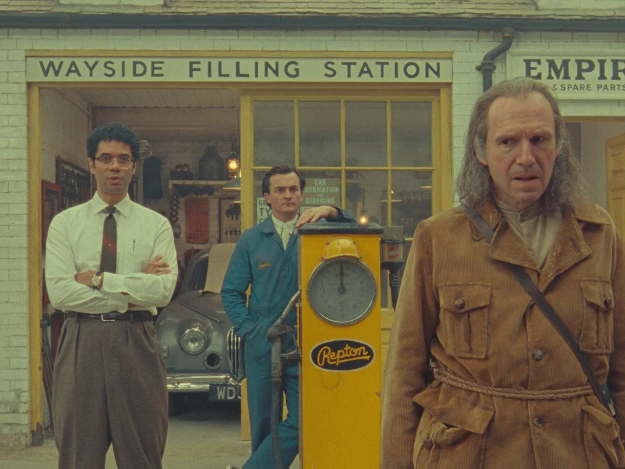 The Rat Catcher (2023) movie still: fullbody — A 1950s filling station facade with a yellow Repton gas pump in front. Three men stand in a line…