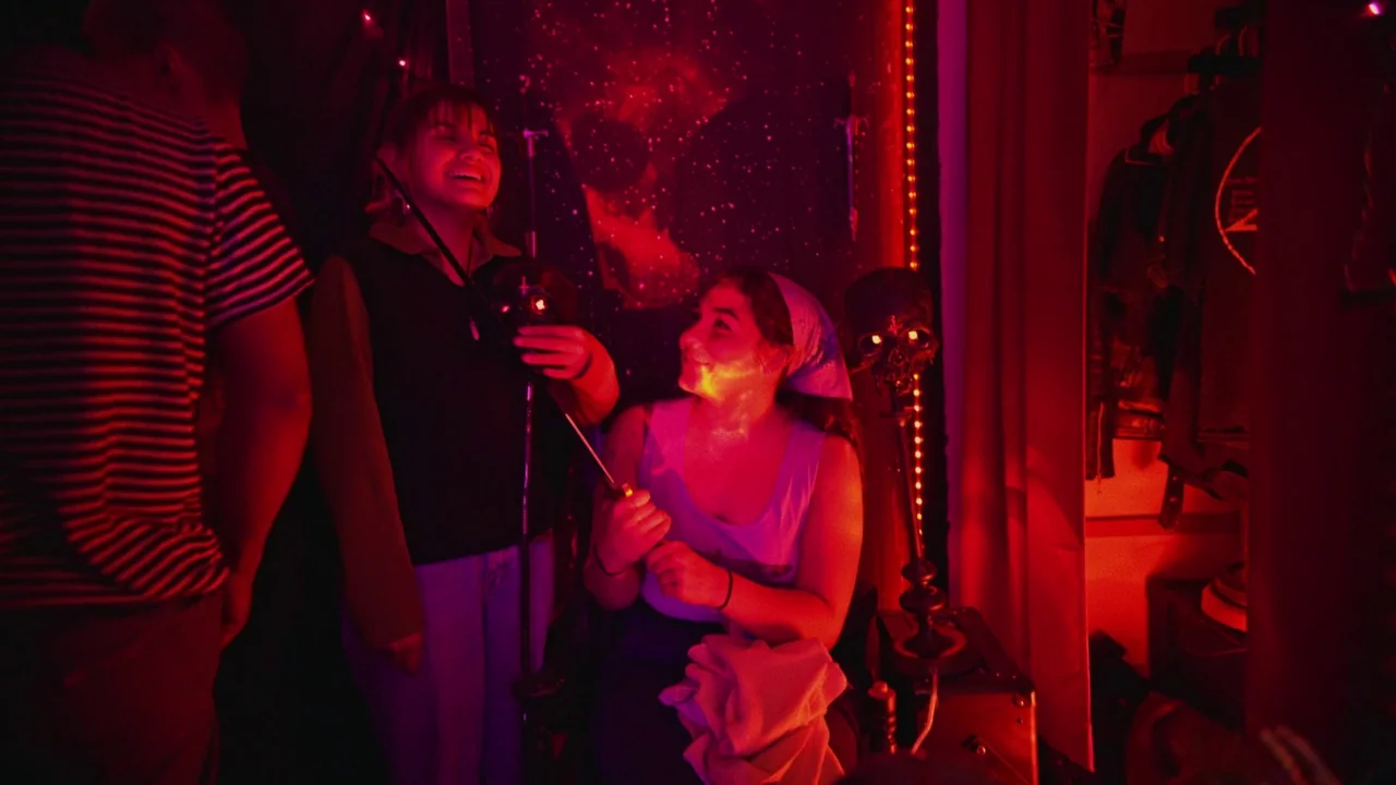 Gasoline Rainbow (2023) movie still: medium — A dimly lit room with walls decorated with a cosmic space pattern tapestry. Two young women laugh…