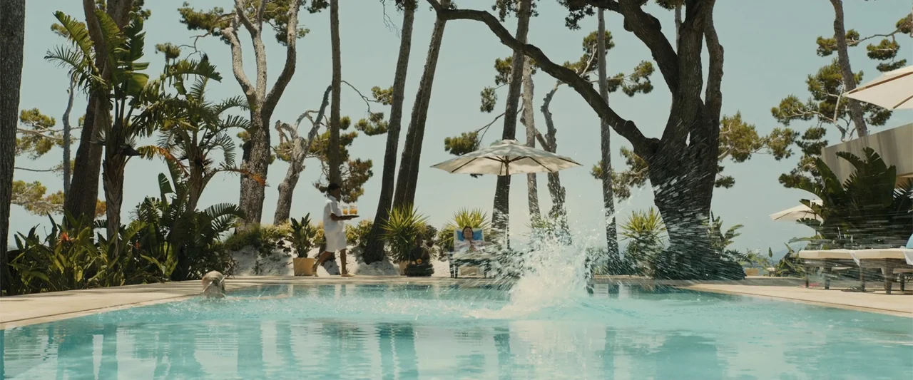 Me Before You (2016) movie still: wide — A sunny pool area features tall pine trees and lounge chairs under umbrellas. A person enters the water…