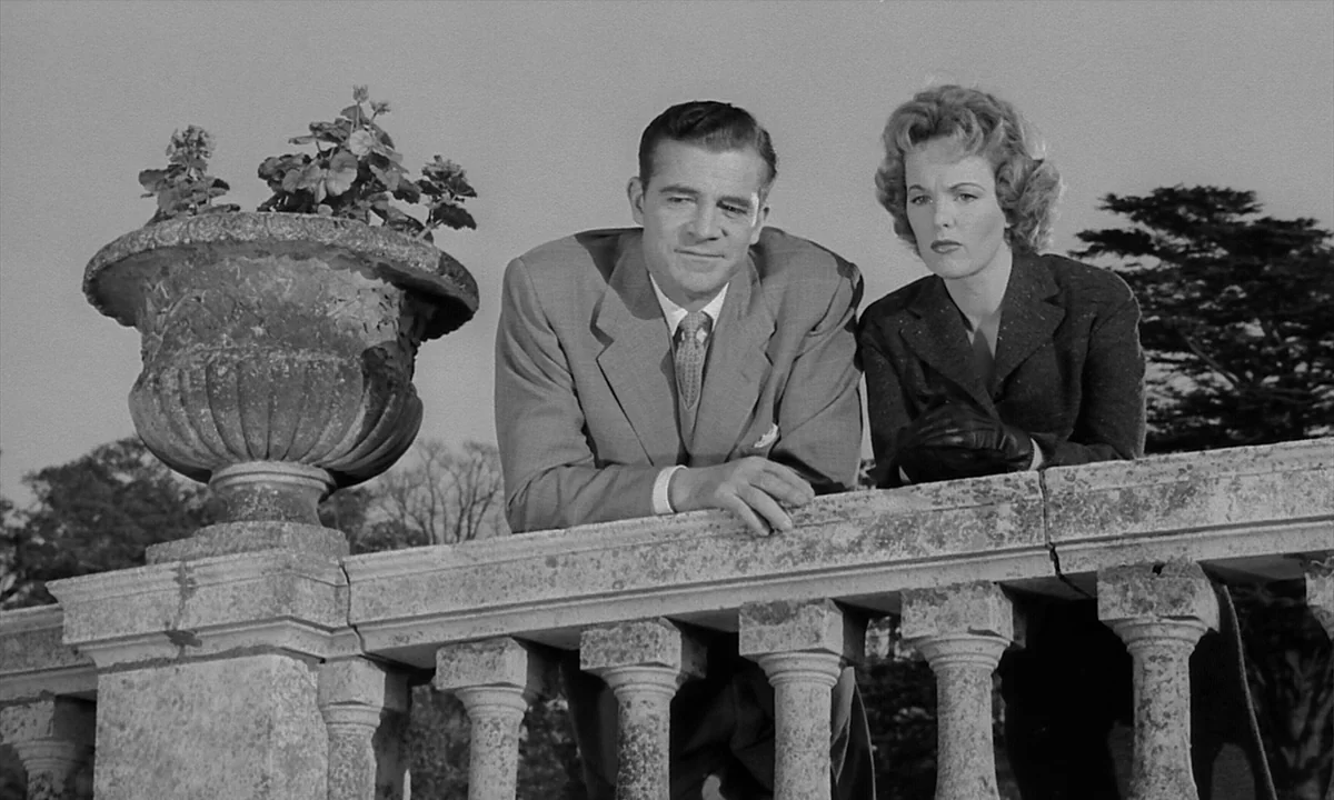 Night Of The Demon (1957) movie still: medium — A man and a woman stand together on an outdoor stone terrace. They lean against a weathered…