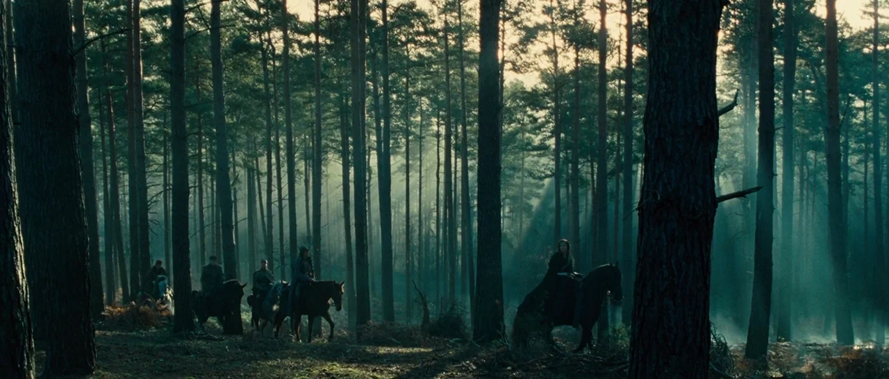 Wonder Woman (2017) movie still: wide — A dense pine wood features tall, vertical trunks and a misty, ground-level atmosphere. A group of riders on…