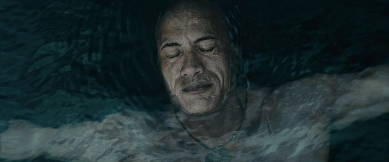 Black Adam (2022) movie still: closeup — A dark body of water fills the frame with rippling surface textures. A man lies horizontally under the…