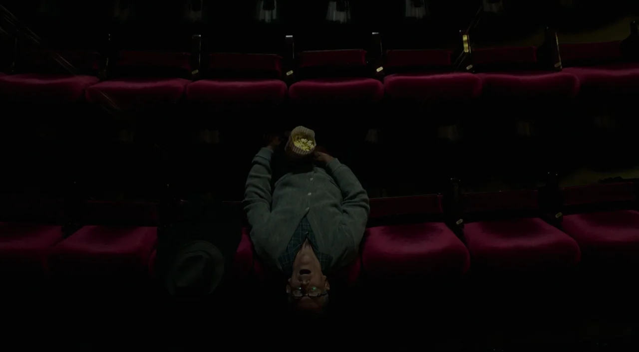 The Shape of Water (2017) movie still: wide — Rows of empty red velvet seats in a dark movie theater are visible. An elderly man lies across the…
