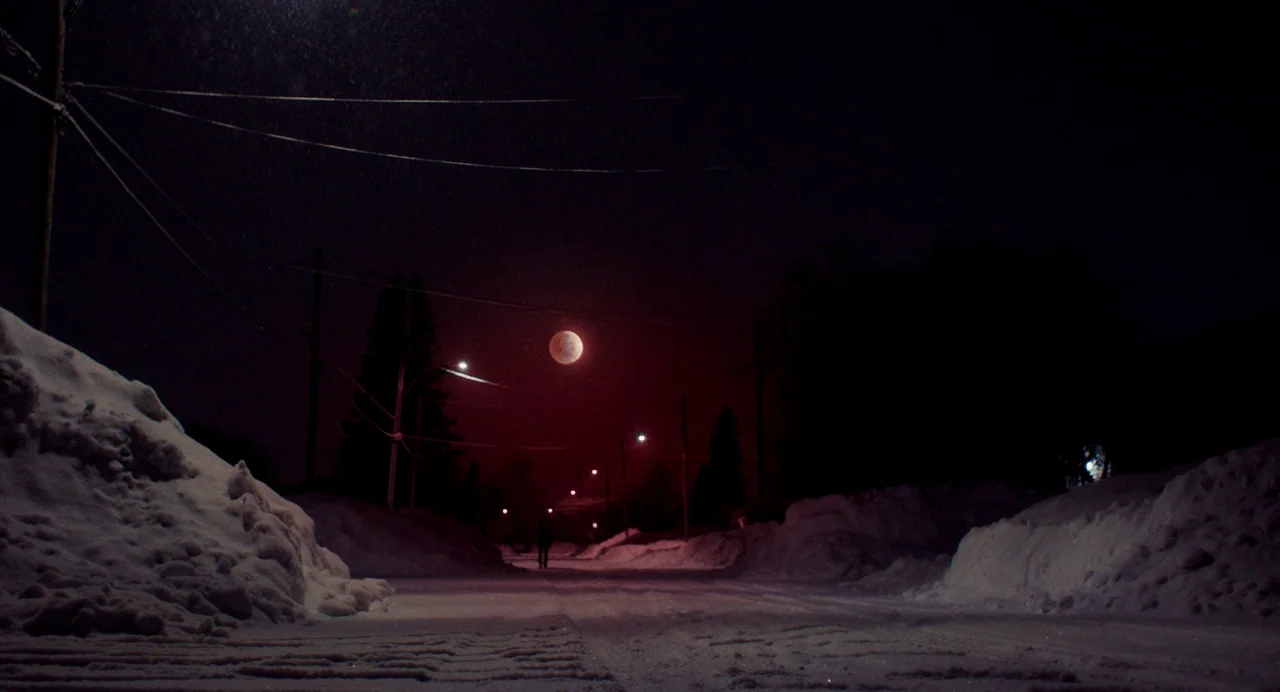 My Animal (2023) movie still: wide — A snow-covered road at night is flanked by high piles of plowed snow. A lone figure walks into the distance…