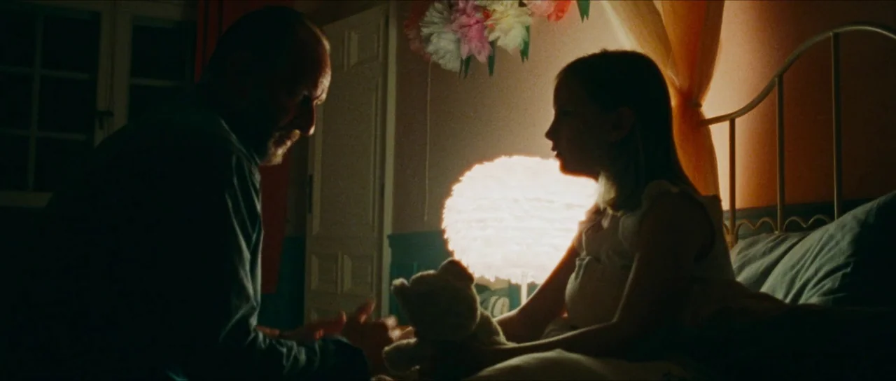 Inexorable (2021) movie still: medium — A child's room is illuminated by a glowing, round feathered lamp placed between two people. A man sits on…