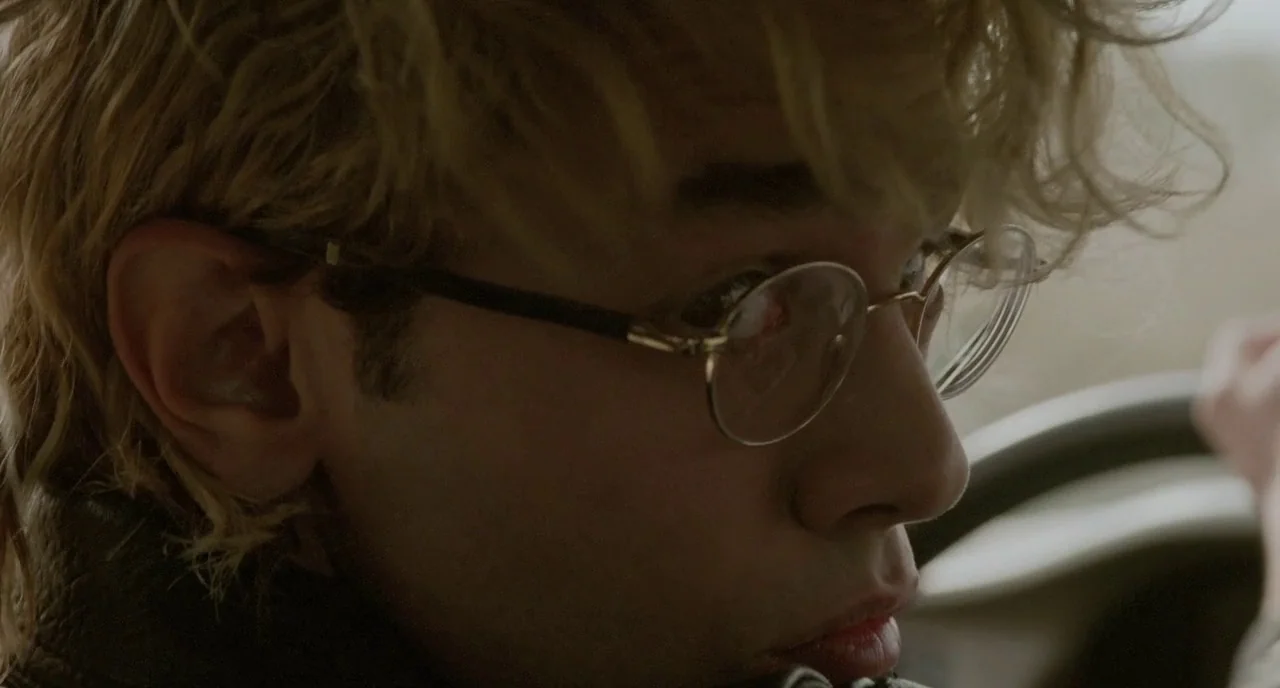 Tom at the Farm (2013) movie still: closeup — A man drives a vehicle while wearing round metal-framed glasses. His messy blond hair obscures his…
