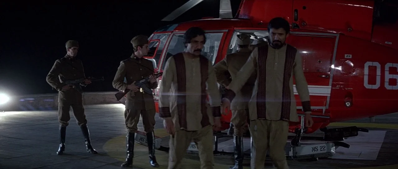 Octopussy (1983) movie still: fullbody — A concrete helipad at night features a large red helicopter parked near the edge. Two men in traditional…