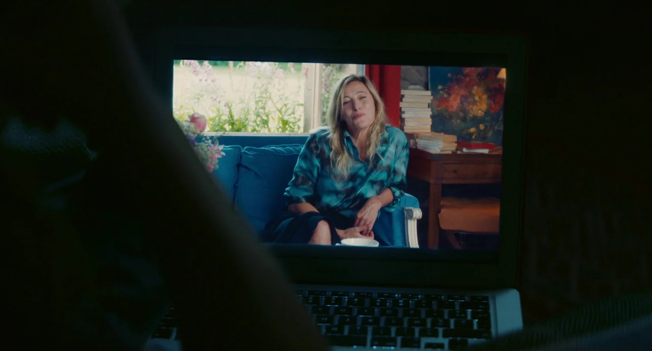 Anaïs in Love (2021) movie still: medium — A woman appears on the screen of a laptop placed in a dark, out-of-focus room. The woman sits on a blue…