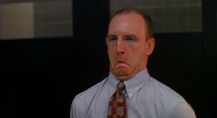 Schizopolis movie still: closeup — A man stands in an office with dark wall panels. He makes a grimace face towards the camera.