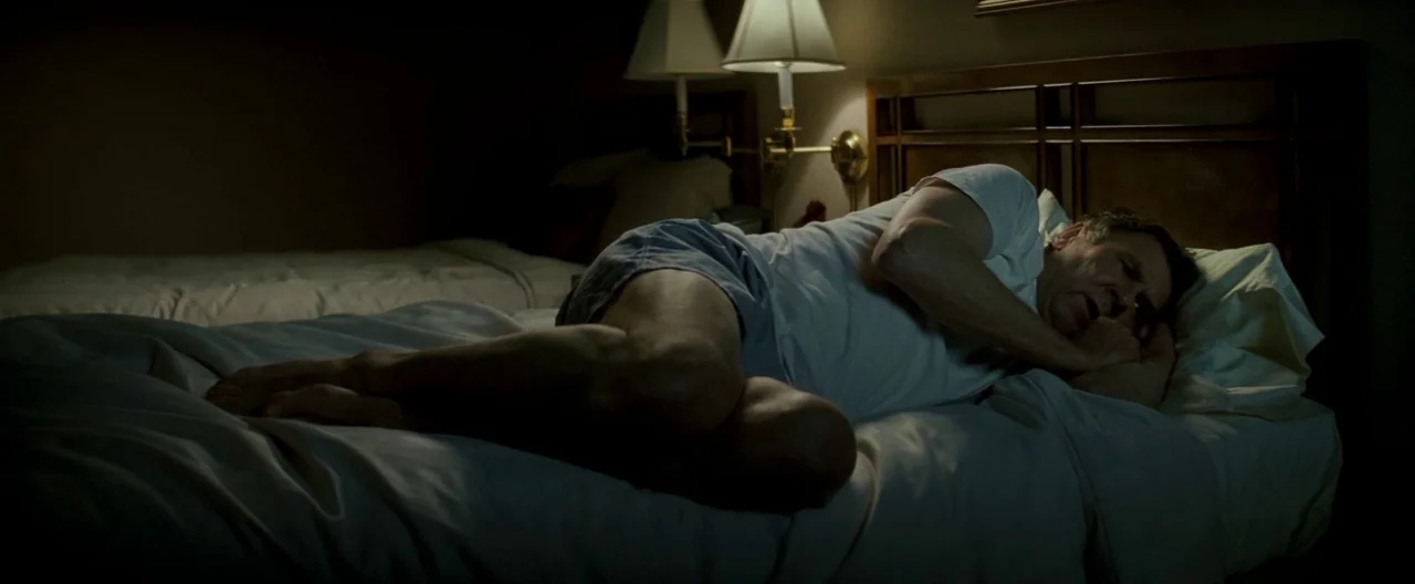 Michael Clayton (2007) movie still: medium — A dimly lit hotel room features two beds with plain white linens and bedding. An older man lies curled…