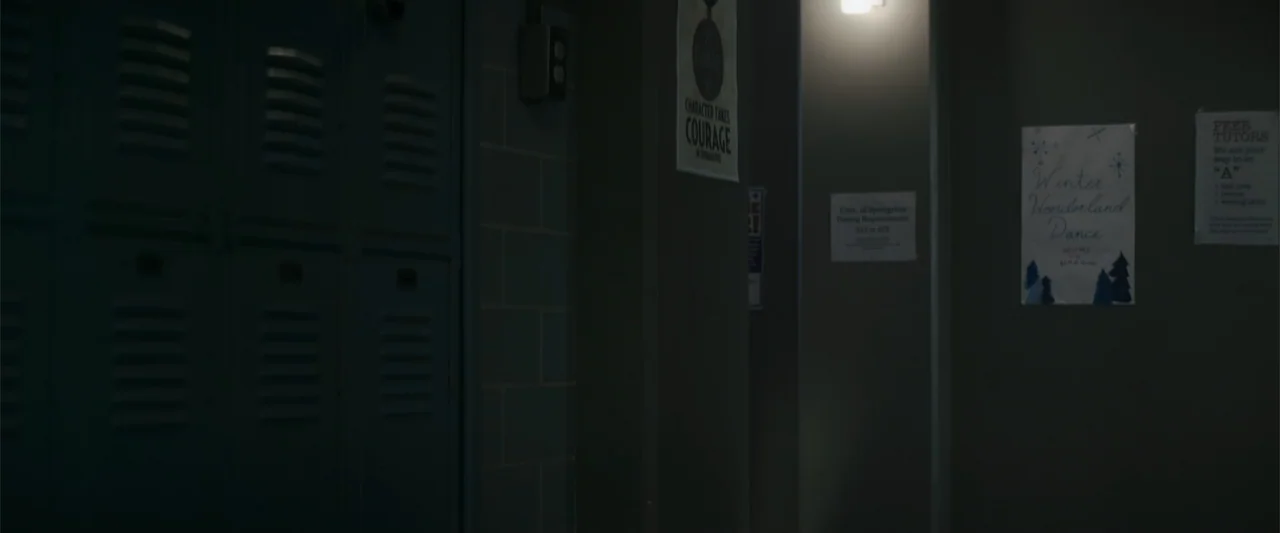 A Christmas Mystery (2022) movie still: wide — A dark school hallway with metal lockers and paper flyers on the wall. The space is dimly lit by a…