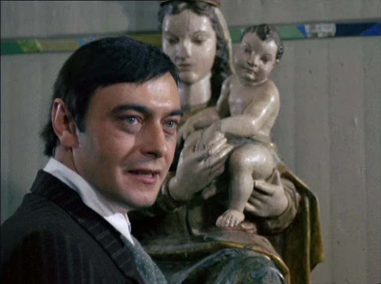 Satan’s Brew (1976) movie still: medium — A man stands in an interior space next to a religious statue. The carved wooden figure depicts a woman…