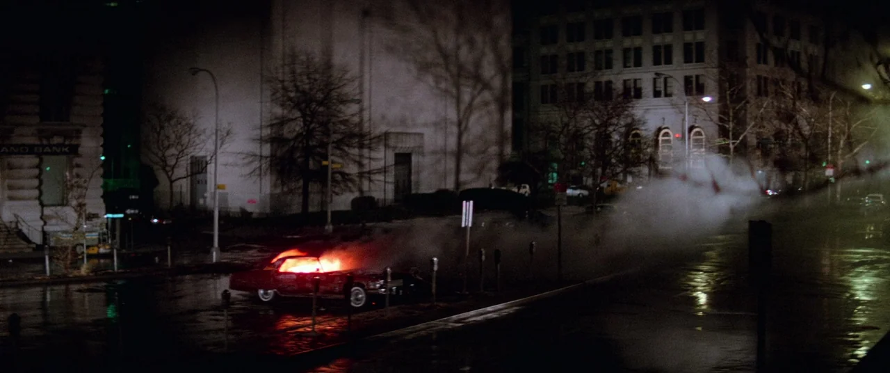 Vigilante (1982) movie still: wide — A rain-slicked city street features a red car engulfed in flames near a bank. Smoke drifts across the empty…