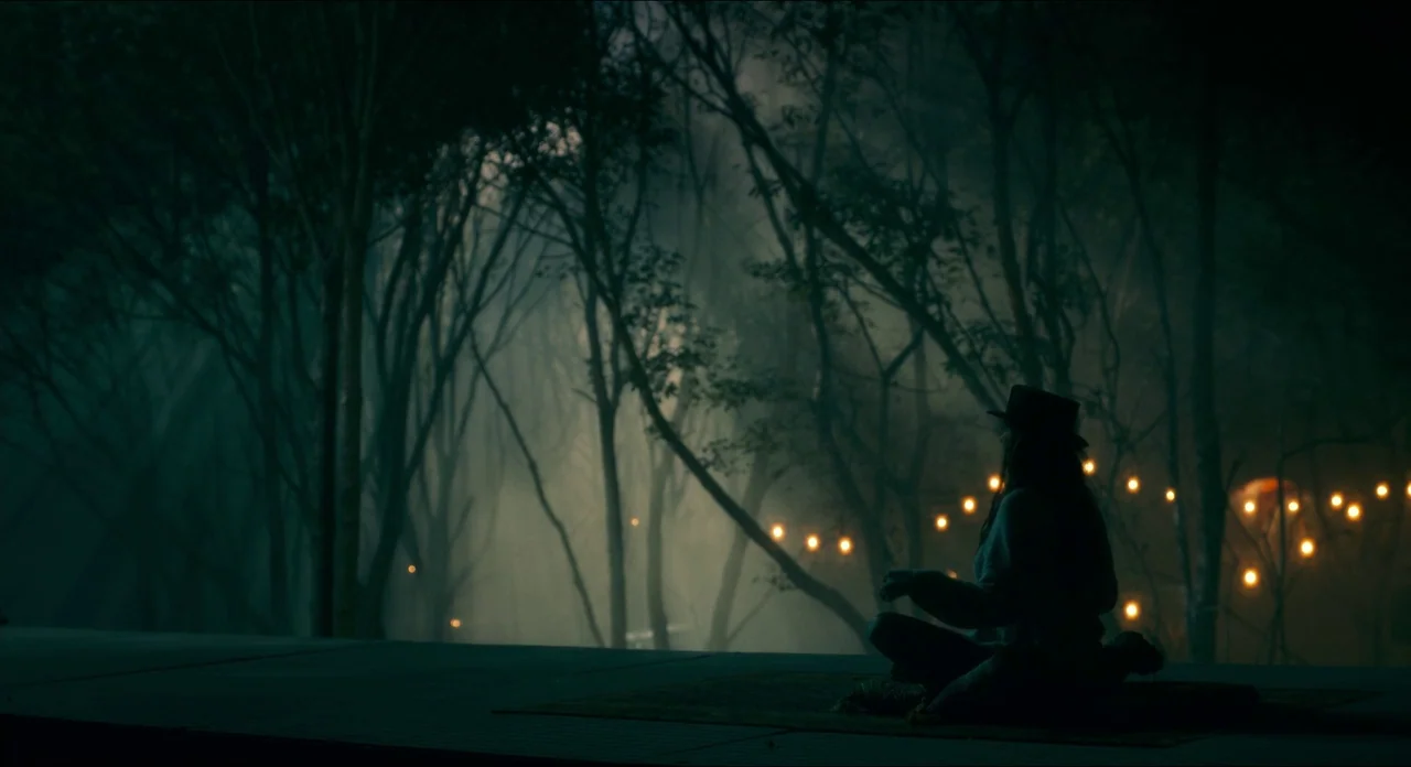 Doctor Sleep movie still: wide — A dark, misty woodland area at night with tall trees and thick fog. A lone person sits on a mat on a raised wooden…