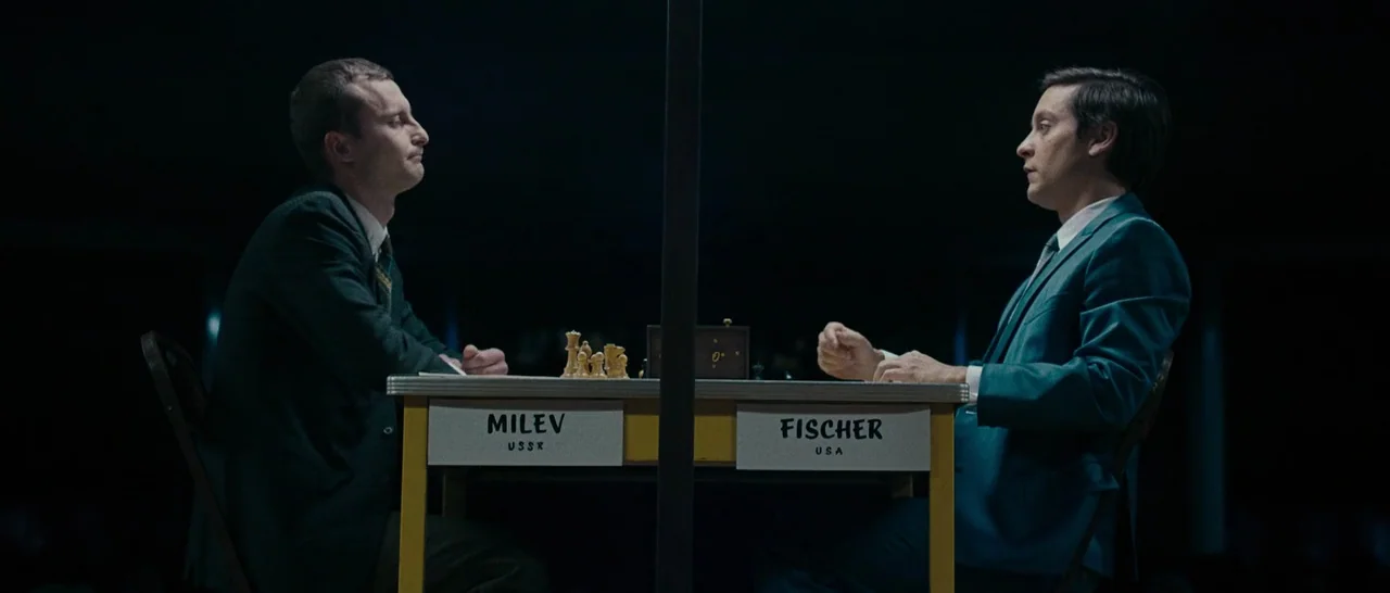 Pawn Sacrifice (2014) movie still: medium — A dark, sparse tournament venue with a single table positioned between two seated men. The table has…