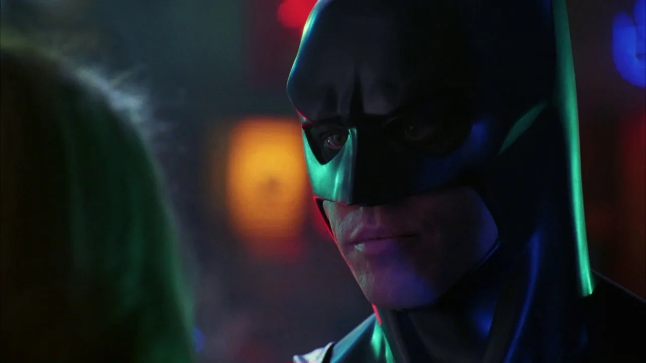 Batman Forever (1995) movie still: closeup — A dimly lit club interior with blurred colorful light bokeh in the background. Batman wearing a black…