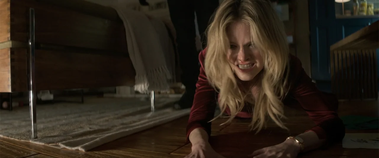 Drop (2025) movie still: medium — A low-angle view captures a person on the floor of a dimly lit room. The woman crawls across hardwood planks while…