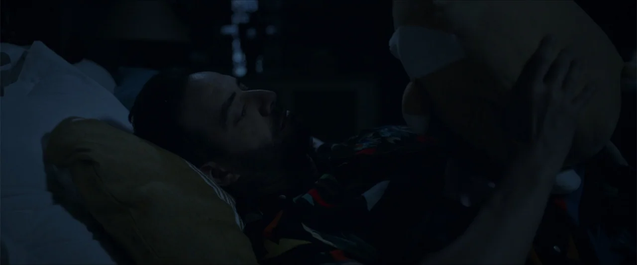 An Endless Wedding (2025) movie still: closeup — A dark bedroom with white pillows and bedding visible under dim, low-key lighting. A man with a…
