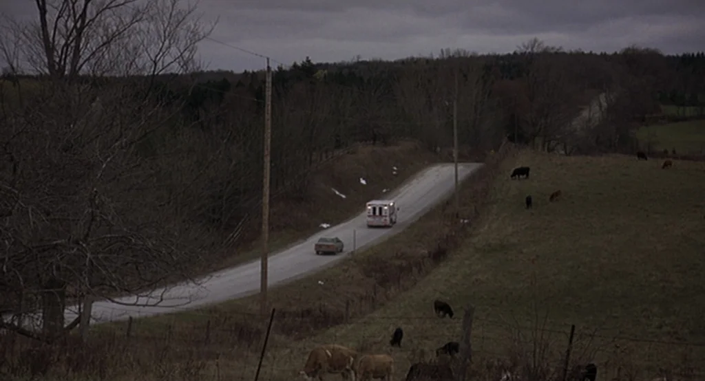 Lars And The Real Girl (2007) movie still: wide — A quiet rural two-lane road stretches through a hilly landscape under overcast grey skies. An…