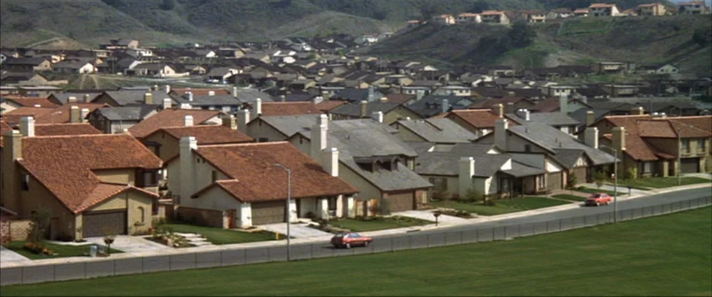 Poltergeist movie still: establishing — A sprawling suburban neighborhood features dense rows of uniform houses with brown tiled roofs. Two small…