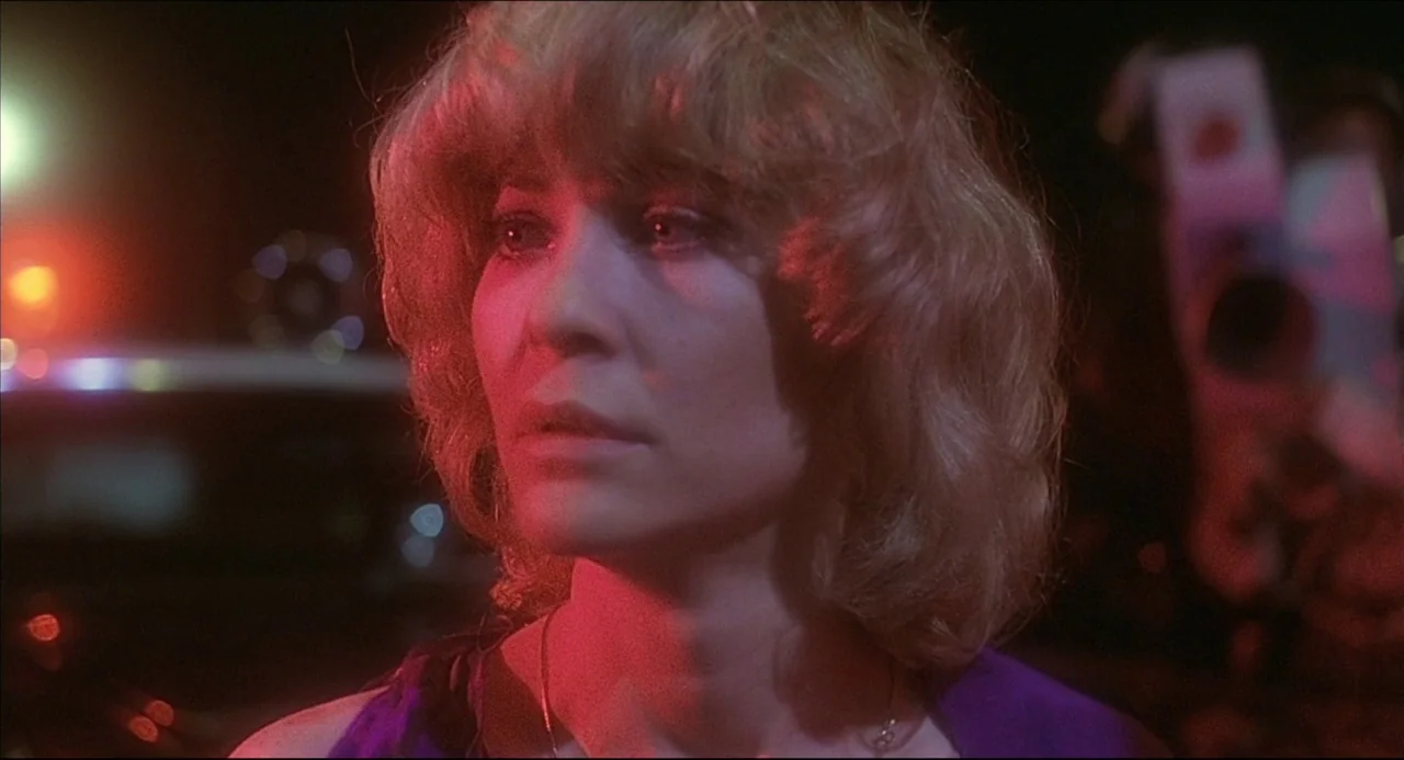The Howling (1981) movie still: closeup — A dark parking lot environment at night with blurred colored lights in the background. A woman with blonde…
