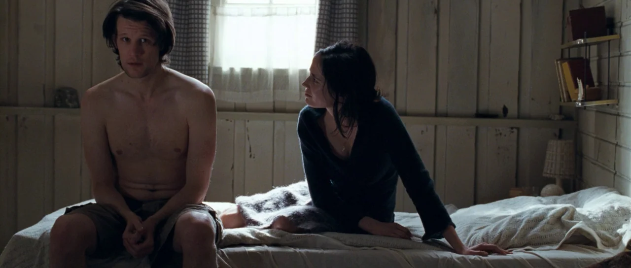 Womb (2010) movie still: medium — A spartan bedroom features wood-paneled walls and a window with a simple curtain. A shirtless man sits on the side…