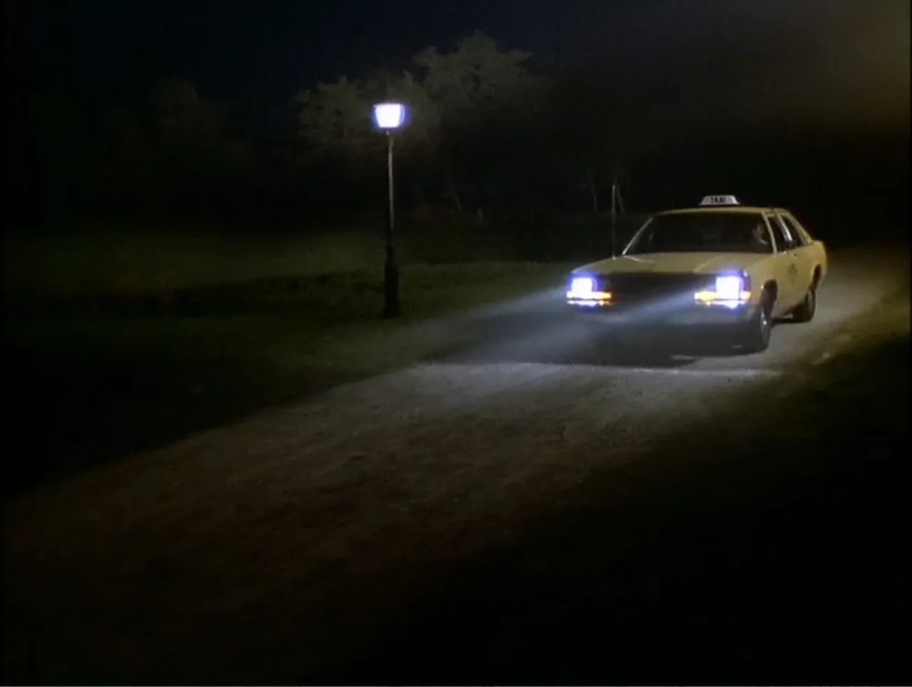 At the Midnight Hour (1995) movie still: wide — A yellow taxi drives down an unpaved road at night in a dark park. The vehicle headlights illuminate…