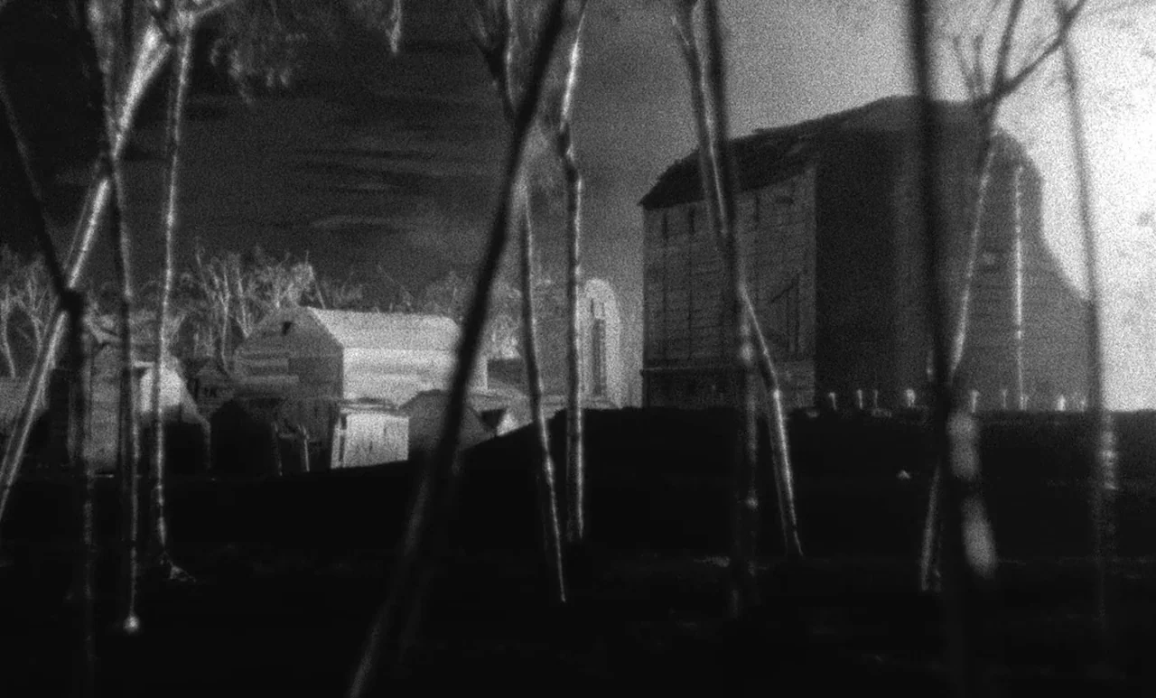 Go Down Death movie still: establishing — A dark rural village features several wooden structures under a night sky. Tall thin sticks or trees in…