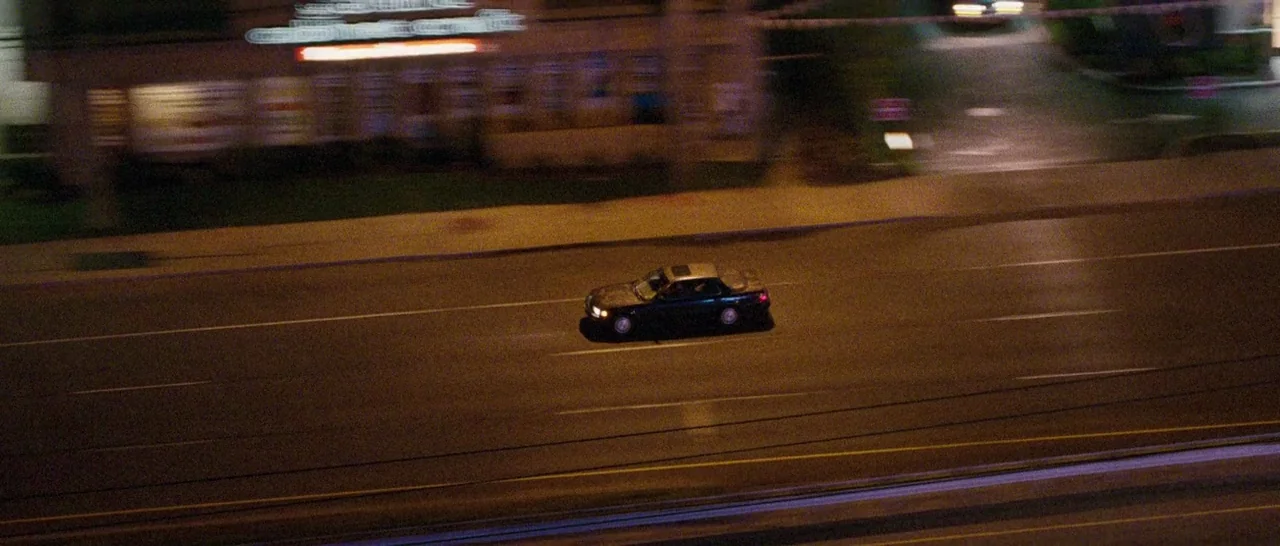 Good Time (2017) movie still: wide — A dark multi-lane highway is flanked by blurry commercial buildings at night. A lone dark sedan drives…