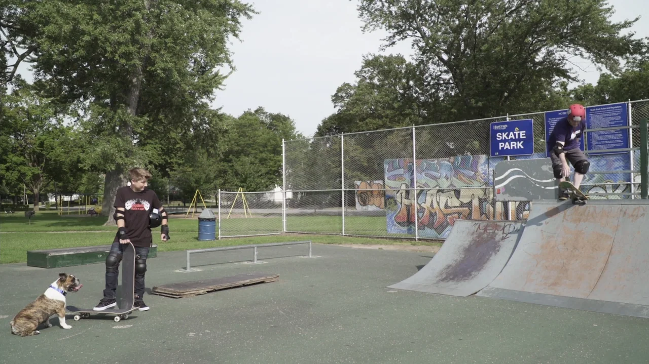 Sk8 Dawg (2018) movie still: wide — A public outdoor recreation area features a metal skate ramp and chain-link fencing. One teenager stands on the…